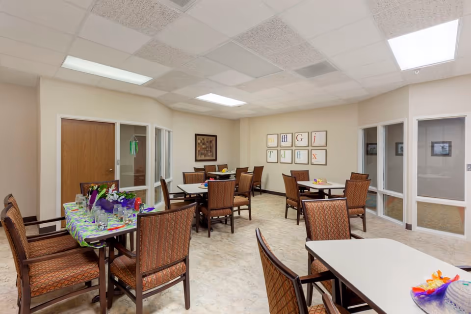 Communal dining/activity room with multiple tables and chairs, colorful table decorations, and framed wall art.