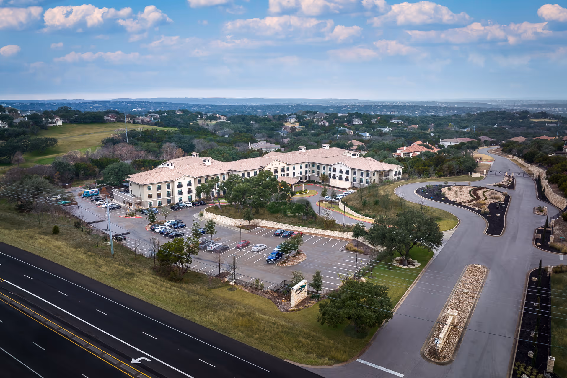 Aerial view of Belmont Village Senior Living Lakeway facility showing a large, two-story building with a beige exterior and a tiled roof. The building is surrounded by a parking lot with several cars and landscaped areas with trees and shrubs. The facility is located near a road with multiple lanes and is set against a backdrop of rolling hills and scattered houses under a partly cloudy sky.
