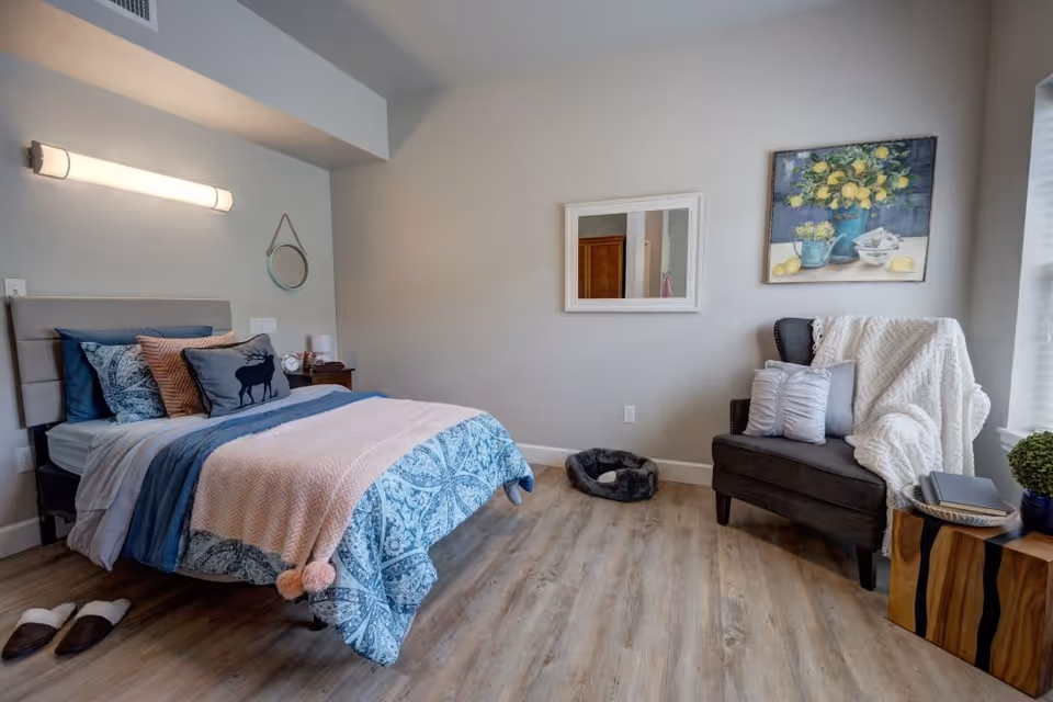 A cozy bedroom with a neatly made bed featuring blue and pink bedding, decorative pillows including one with a moose design, and a padded headboard. To the right, there is a dark gray armchair draped with a white knitted blanket and adorned with pillows. A small wooden side table next to the chair holds a book and a small plant. The room has light wood flooring, a wall-mounted light fixture above the bed, a round mirror hanging on the wall, a rectangular mirror, and a painting of lemons in a blue vase. Slippers are placed on the floor near the bed.