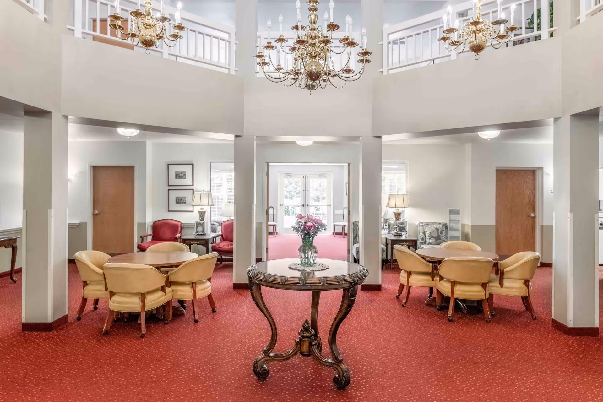 Interior view of a senior living community common area with red carpet flooring, a round wooden table with a vase of flowers in the center, two seating areas with round tables and beige chairs, red and patterned armchairs, and brass chandeliers hanging from the ceiling.