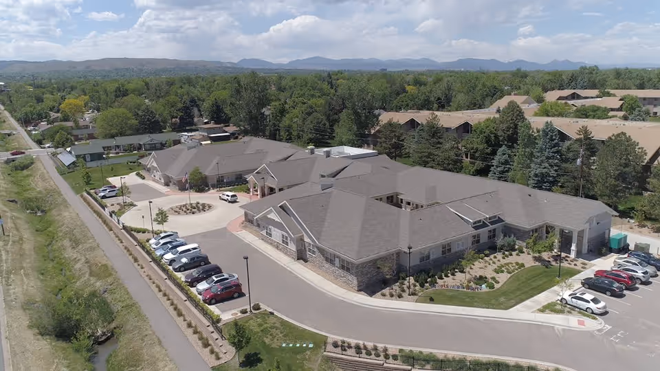 Aerial exterior view of a single-story memory care building with a curved driveway, parked cars, and surrounding trees and landscaping.