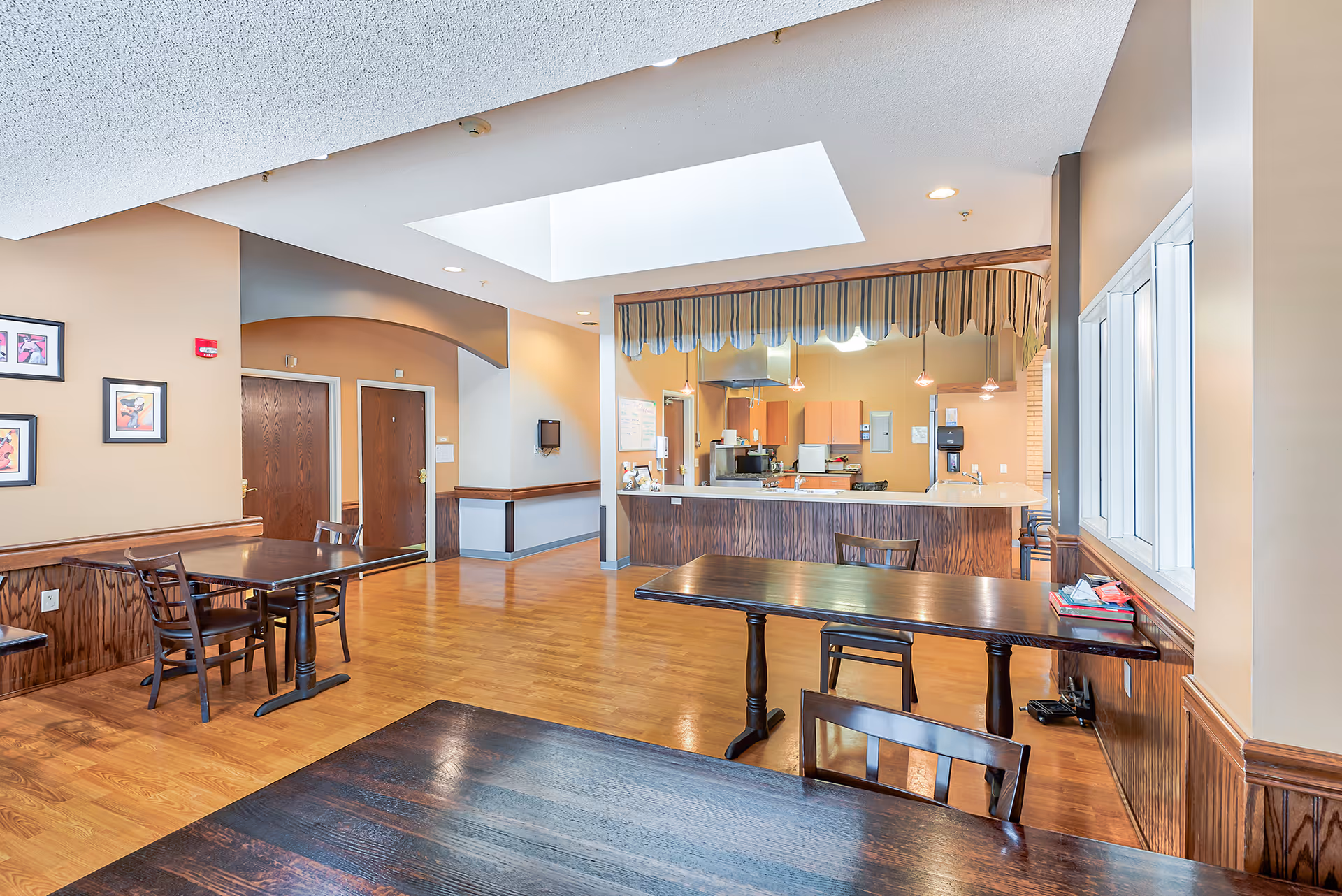 Interior view of a senior living facility dining area with wooden tables and chairs, a kitchen counter with a striped valance above, light wood flooring, beige walls, and a skylight in the ceiling providing natural light.