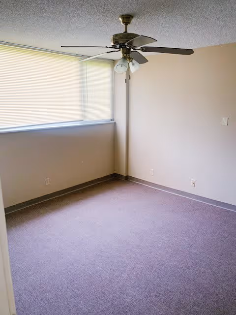 Empty room with beige walls, a ceiling fan with lights, a large window with closed blinds, and carpeted floor.