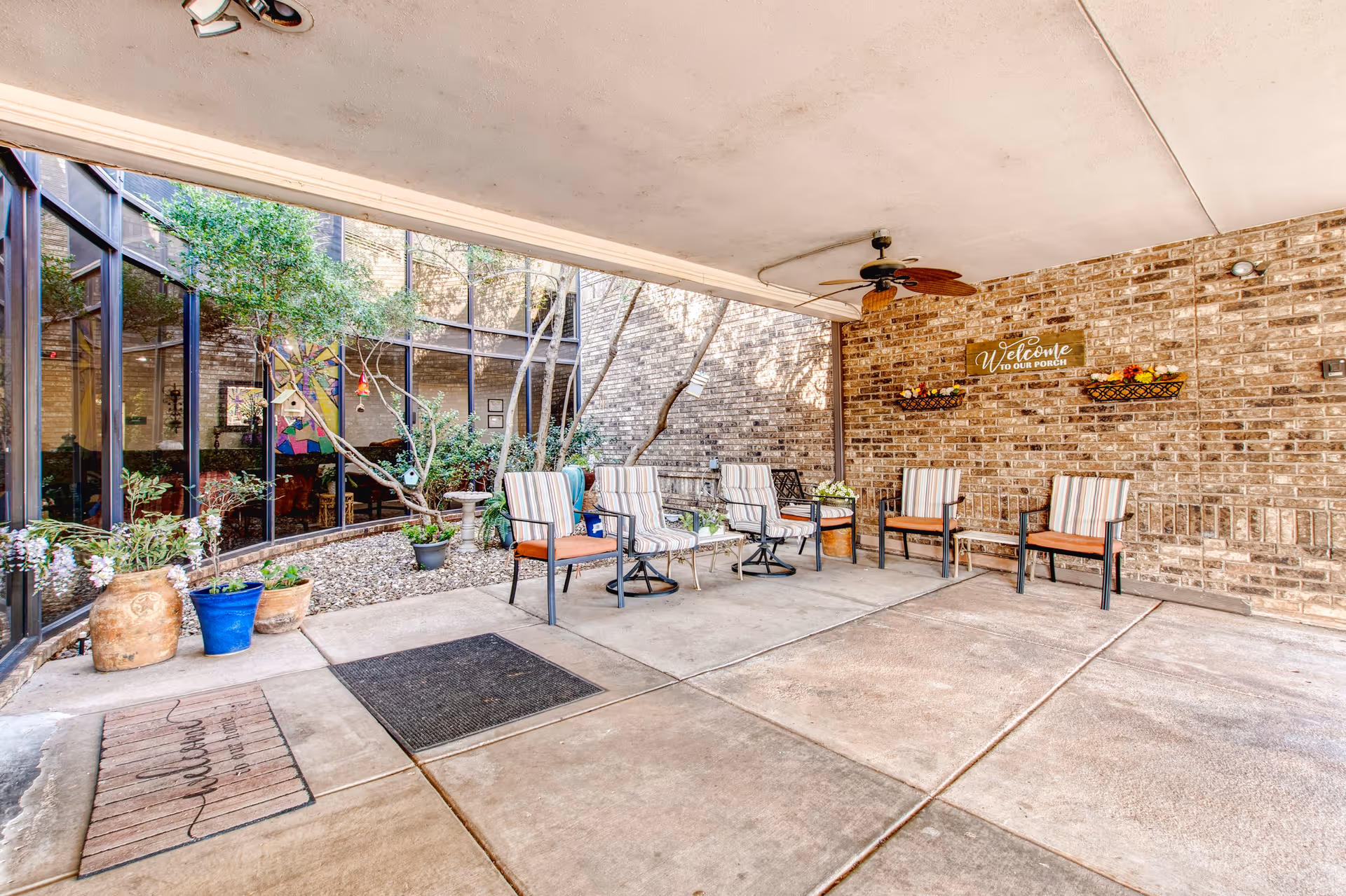 Covered outdoor patio area with several cushioned chairs arranged around a small table. The space features a brick wall with a decorative sign that reads 'Welcome to our porch' and two wall-mounted baskets with flowers. There are potted plants and small trees along the glass windows on the left side, and a ceiling fan is mounted above.