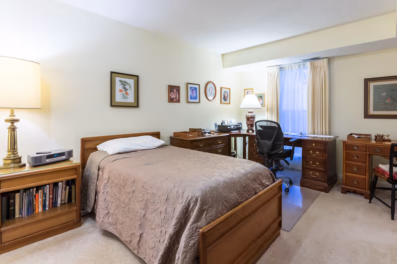 A cozy bedroom with a single bed covered in a beige quilt, a wooden nightstand with a lamp and books, and a wooden desk with an office chair near a window with cream-colored curtains. Several framed pictures hang on the light-colored walls.