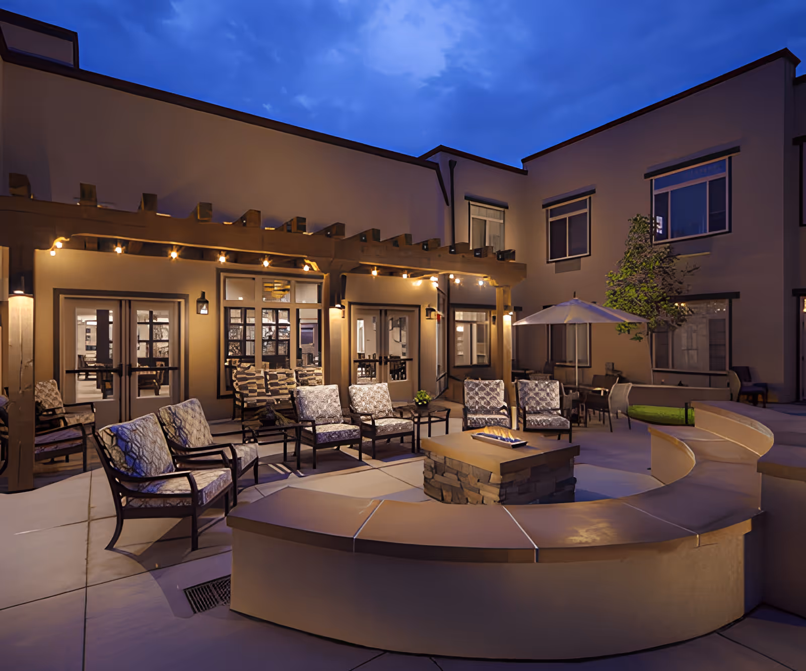 Outdoor courtyard area of MorningStar Assisted Living & Memory Care of Rio Rancho at dusk, featuring cushioned chairs arranged around a stone fire pit, a curved bench, a pergola with string lights, and a patio umbrella near a small tree, all surrounded by the building's exterior walls with windows and doors.