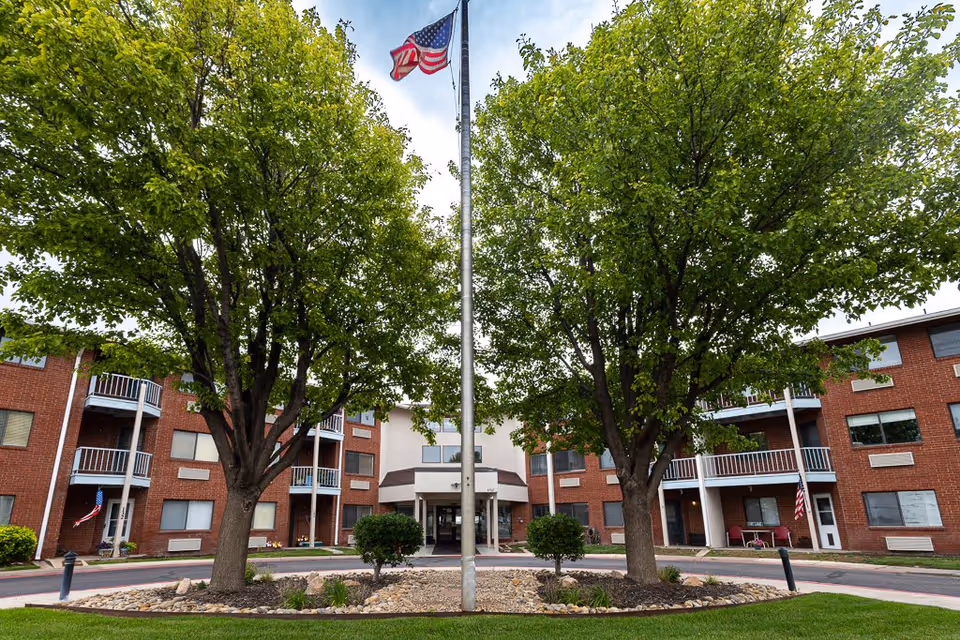Front entrance of a red-brick senior living building with two large trees and a flagpole flying an American flag.