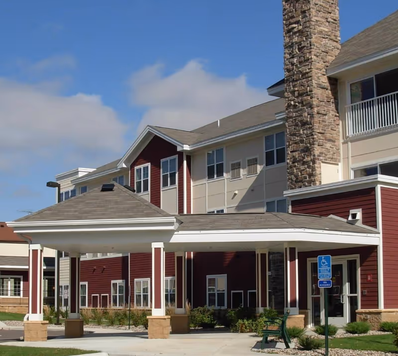 Exterior view of a senior living facility named The Villa with a covered entrance supported by white columns, red and beige siding, multiple windows, a stone chimney, a bench, and a handicap parking sign in front.