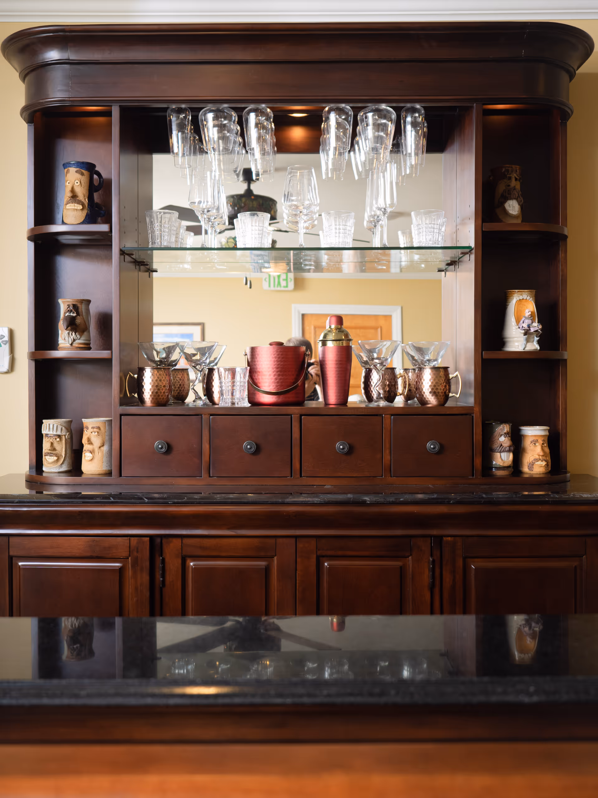 A dark wooden cabinet with glass shelves displaying various glassware including wine glasses, tumblers, and copper mugs. The cabinet has small drawers and open shelves on the sides holding decorative ceramic mugs with faces. A mirror behind the glassware reflects part of the room including a door and an exit sign.