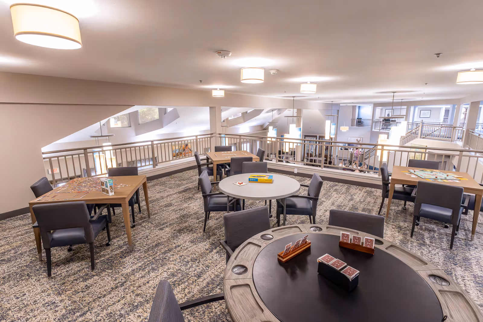 A spacious and well-lit common activity room with multiple tables and chairs arranged for games and socializing. The tables have various board games, puzzles, and playing cards set up. The room has a carpeted floor, modern ceiling lights, and a railing overlooking a lower level.