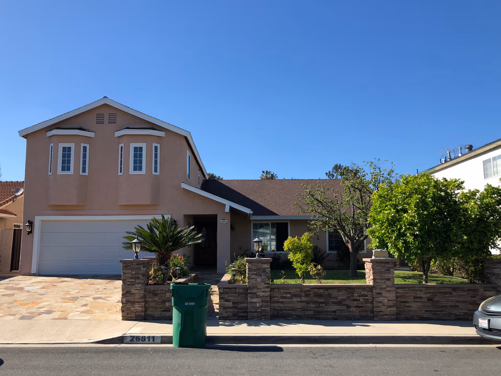 Front exterior of Delightful Living, a beige two-story senior living facility with a garage, stone planters, and a landscaped yard under a clear blue sky.
