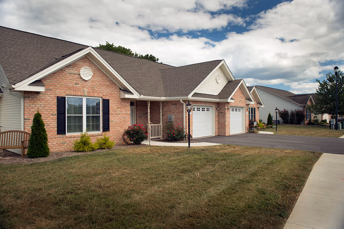 Single-story brick residential units with attached garages, front lawns, and a driveway under a partly cloudy sky.