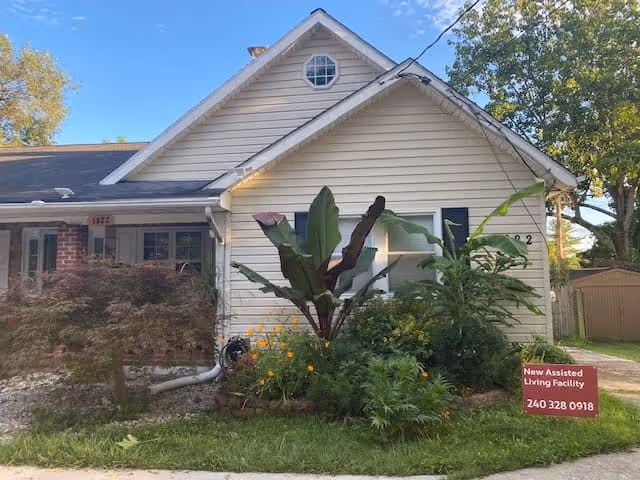 Exterior view of a single-story house with white siding and a dark roof. The front yard has various plants and shrubs, including a large leafy plant in the center. A red sign in the yard reads 'New Assisted Living Facility 240 328 0918'. Trees and a shed are visible in the background under a clear blue sky.