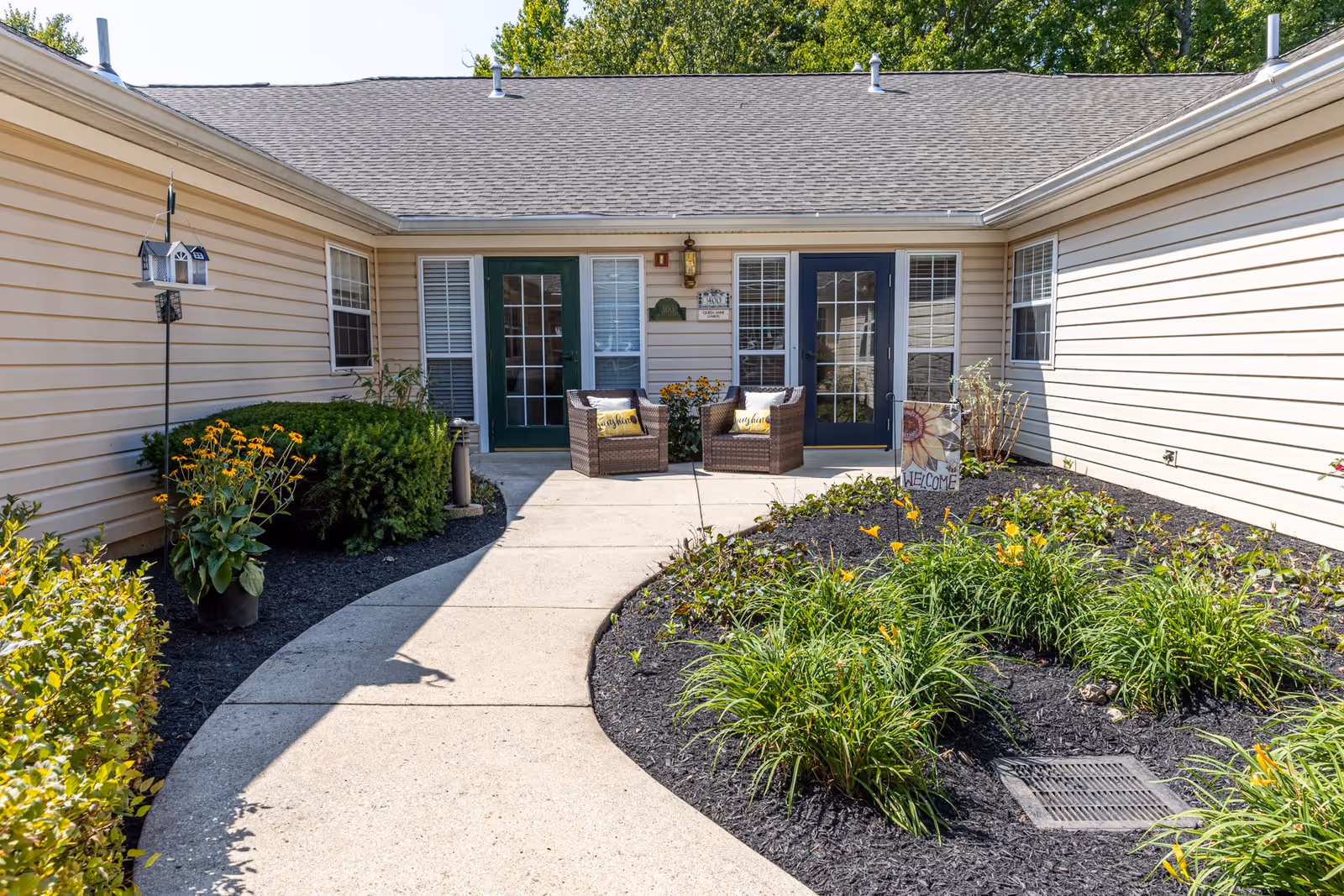 Outdoor courtyard area of a senior living facility with a curved concrete walkway leading to two sets of glass-paneled doors. There are two wicker chairs with yellow pillows placed between the doors, surrounded by flower beds with green plants and yellow flowers. A decorative welcome sign and a birdhouse on a pole are also visible.