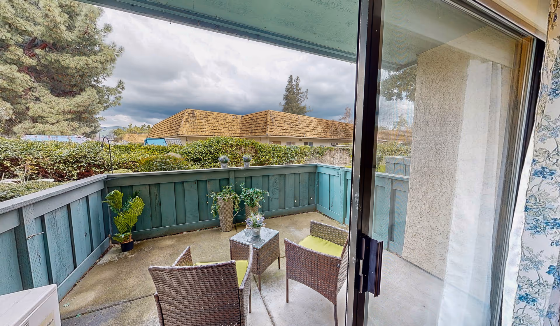 View of a small outdoor patio area with two wicker chairs with green cushions and a small glass-top table between them. There are potted plants along the teal wooden fence surrounding the patio. Beyond the fence, there are bushes, trees, and a building with a brown shingled roof under a cloudy sky. The patio is accessed through a sliding glass door with floral curtains on the right side.