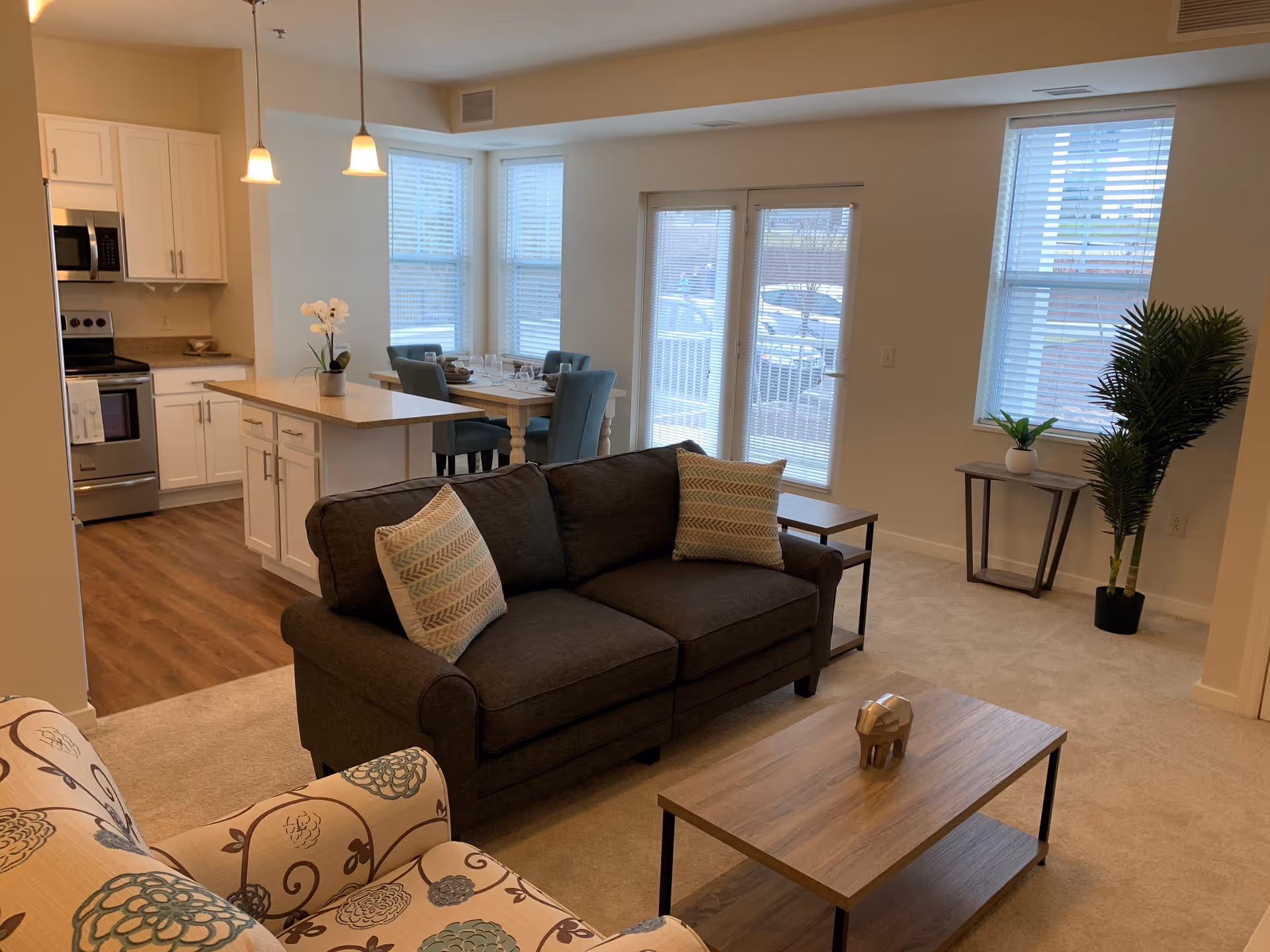 A bright and cozy living room and kitchen area in a senior living facility. The living room features a dark gray sofa with patterned pillows, a floral armchair, a wooden coffee table with a decorative item, and a side table with a plant. The kitchen has white cabinets, stainless steel appliances, and a small island with a potted orchid. A dining table with four chairs is positioned near large windows and glass doors that let in natural light.