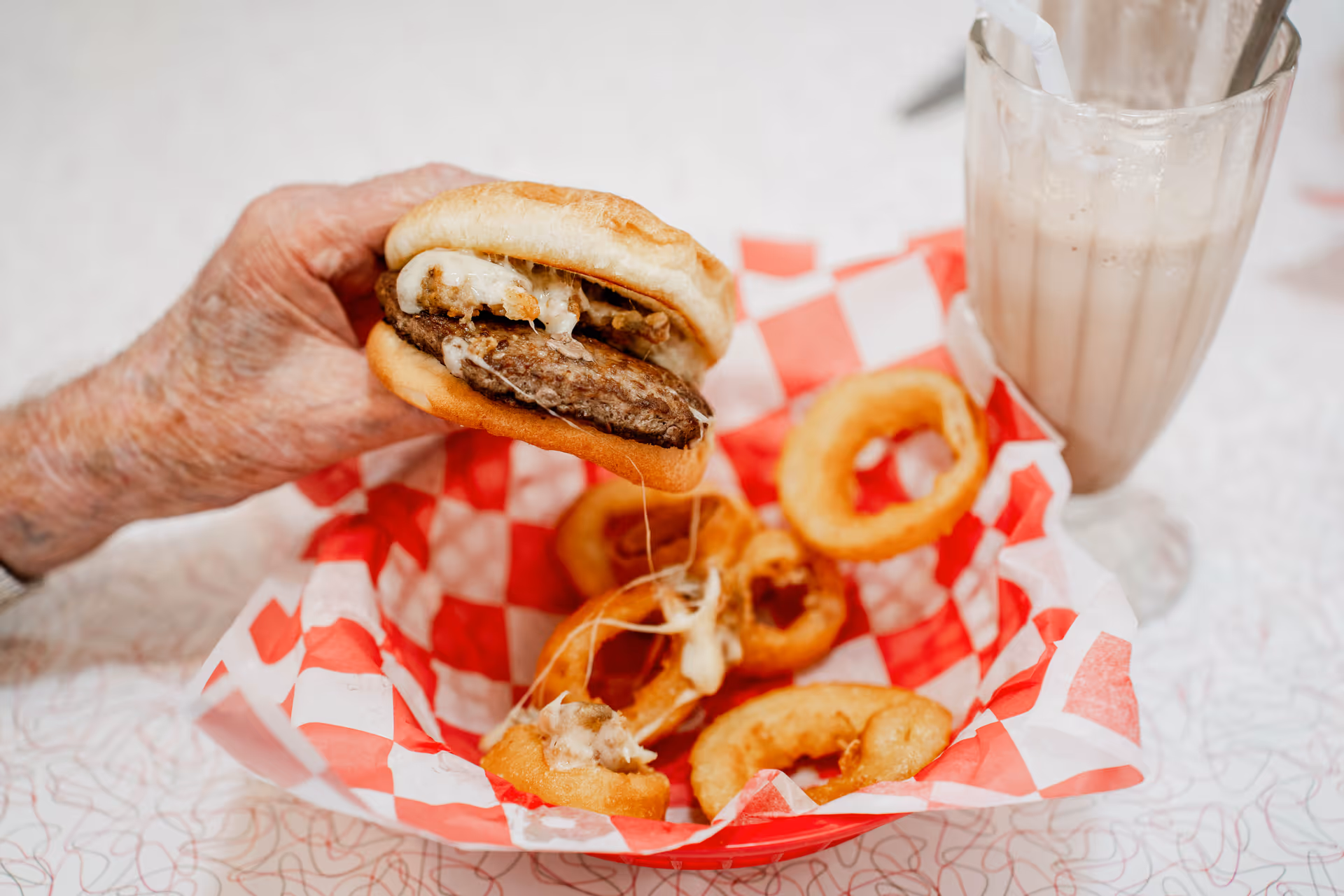An elderly hand holding a cheeseburger above a basket lined with red and white checkered paper containing several onion rings, next to a glass of milkshake with a straw on a white table.
