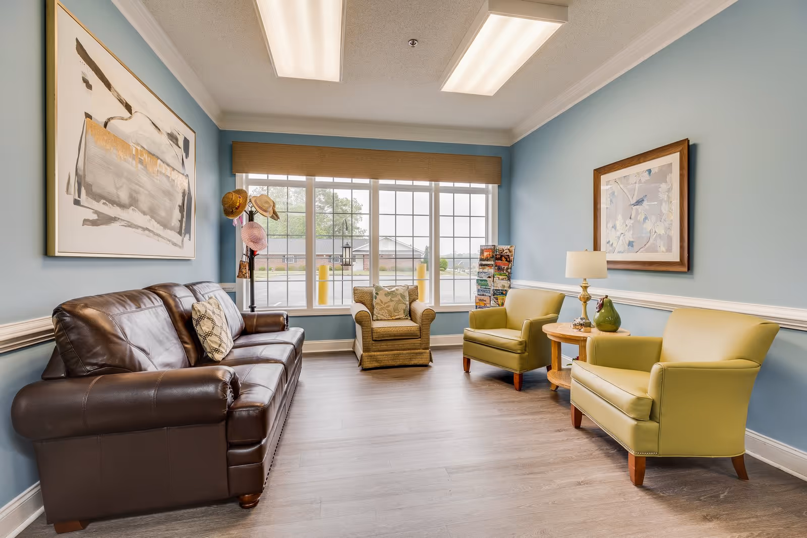 A cozy sitting area in an assisted living facility with a brown leather sofa, two light green armchairs, and a beige armchair near a large window. The walls are painted light blue and decorated with framed artwork. A small wooden side table with a lamp and decorative items is placed between the green armchairs. A coat rack with hats is positioned near the window.
