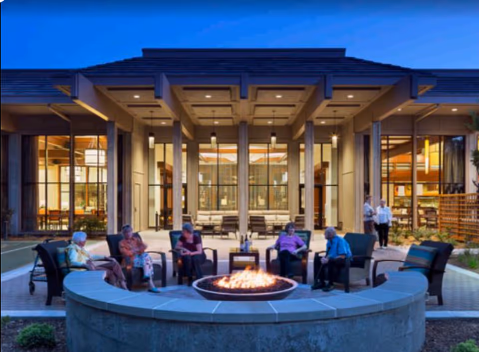 A group of elderly people sitting around a circular outdoor fire pit in front of a modern building with large windows and warm interior lighting during dusk.