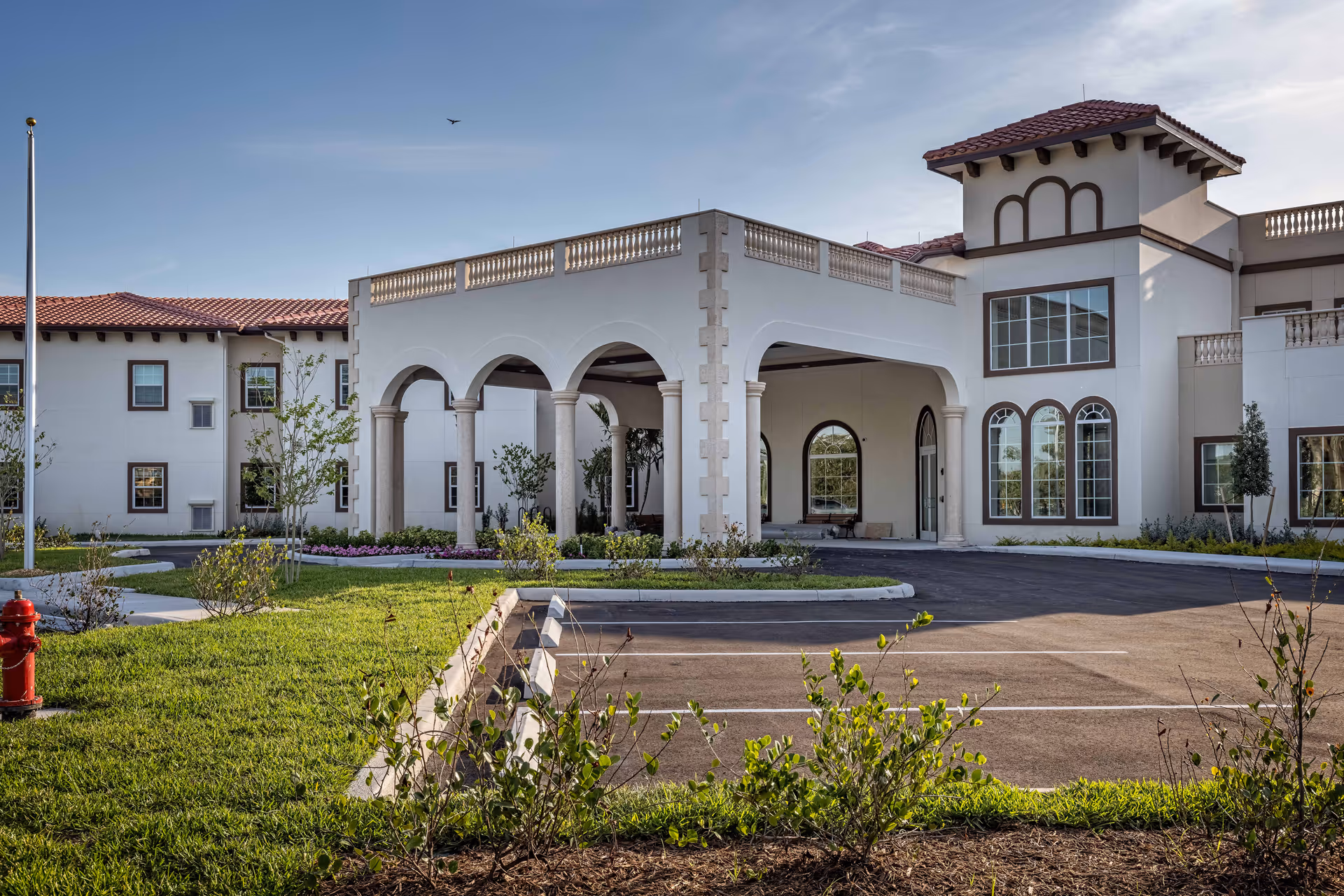 Exterior view of a senior living facility with a covered entrance supported by columns, arched windows, and a red-tiled roof. There is a parking area in front and landscaped greenery surrounding the building under a clear sky.