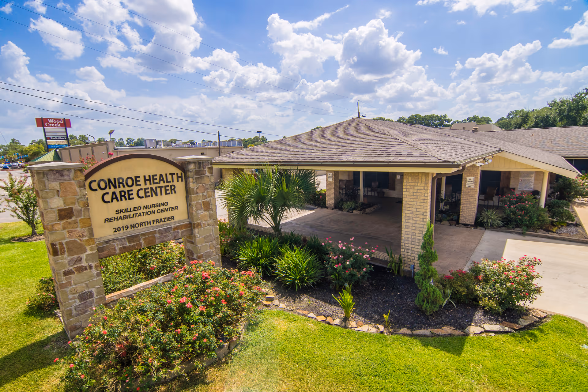 Exterior view of Conroe Health Care Center showing the entrance with a covered driveway, surrounded by well-maintained landscaping including bushes and flowers, under a partly cloudy sky.