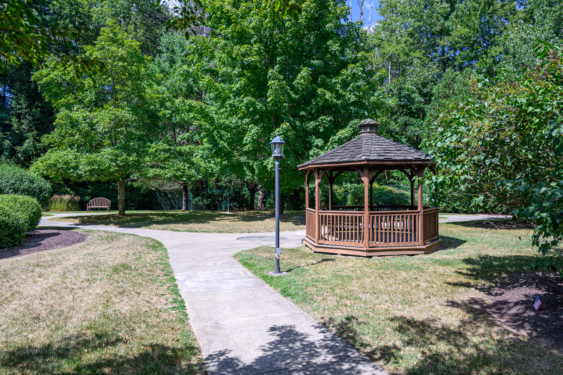 A peaceful outdoor garden area with a wooden gazebo, a lamppost, a curved concrete pathway, green trees, bushes, and a wooden bench in the background.