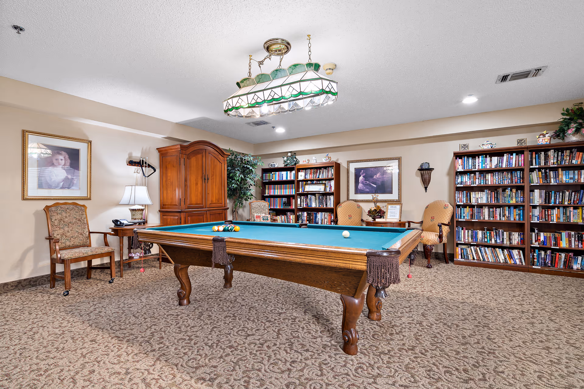 A cozy room with a pool table in the center, surrounded by bookshelves filled with books. There are several upholstered chairs, a wooden cabinet, framed pictures on the walls, a table lamp, and a hanging light fixture above the pool table. The room has carpeted flooring and a warm, inviting atmosphere.