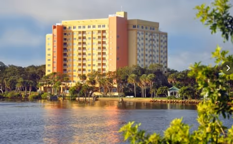 A multi-story residential building with yellow and orange exterior walls situated near a body of water, surrounded by trees and greenery under a partly cloudy sky.