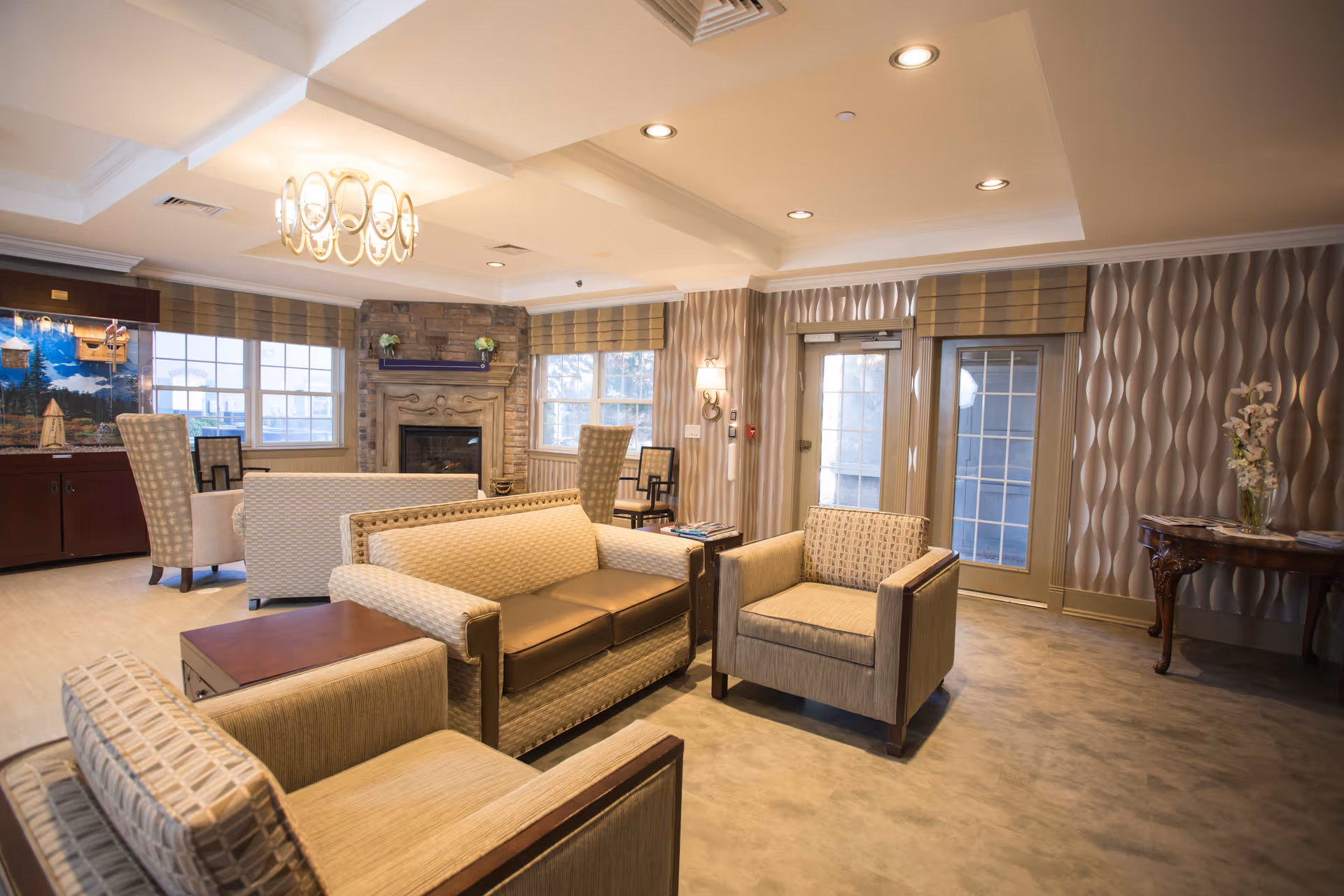 A cozy living room area in a senior living facility with beige upholstered armchairs and a sofa arranged around a wooden coffee table. The room features a stone fireplace with decorative items on the mantle, large windows with blinds, a chandelier, and patterned wallpaper. There is also a wooden side table with a vase of flowers near a glass door.