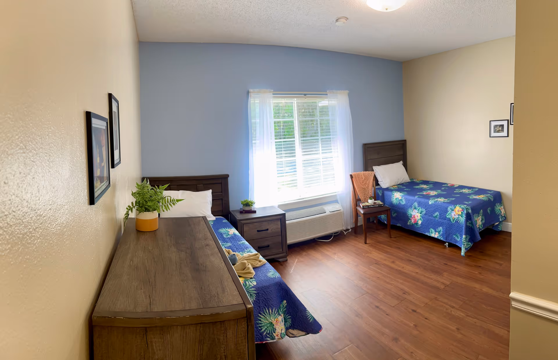 A bright bedroom with two single beds covered in blue floral bedspreads, a wooden dresser with a potted plant, a wooden nightstand between the beds, a wooden chair with a folded blanket and a book, and a window with white sheer curtains letting in natural light.