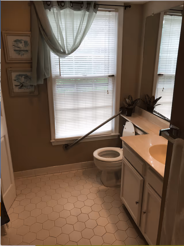 A bathroom with a white toilet, a white vanity with a sink, a large mirror above the sink, and a window with white blinds and a sheer curtain. There is a safety grab bar installed next to the toilet. The floor has hexagonal tiles, and two framed pictures hang on the wall beside the window.