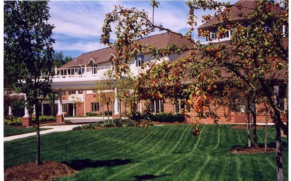 Exterior view of a senior living facility with well-maintained green lawn, trees with some autumn leaves, and a multi-story brick building with balconies under a partly cloudy sky.