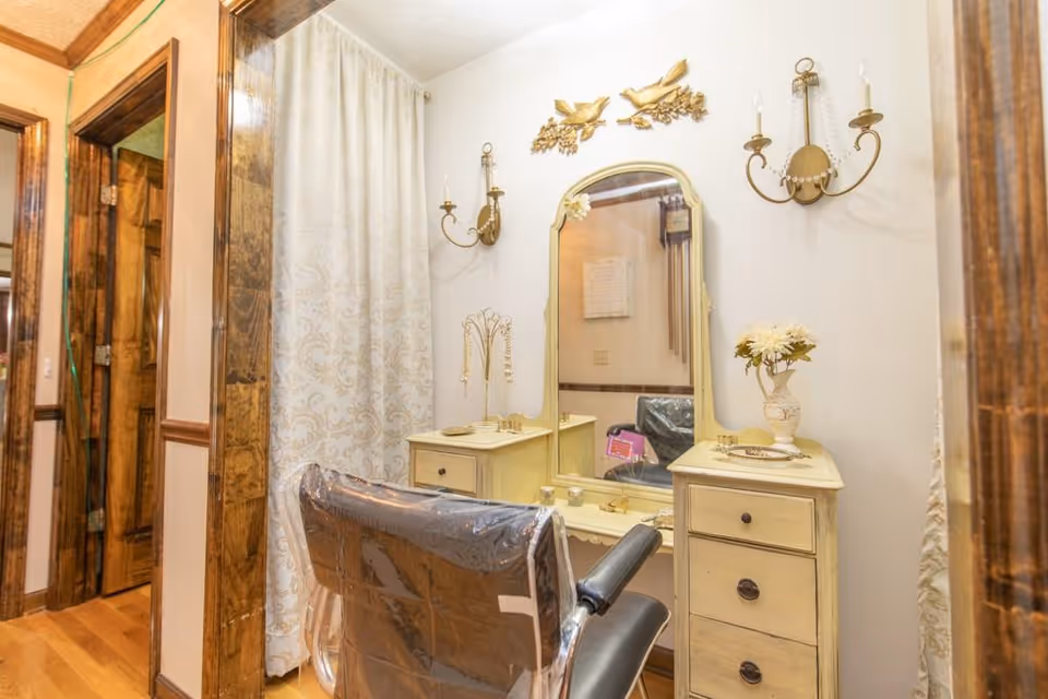 Antique-style dressing vanity with mirror and drawers, a plastic-covered salon chair, and decorative wall sconces.