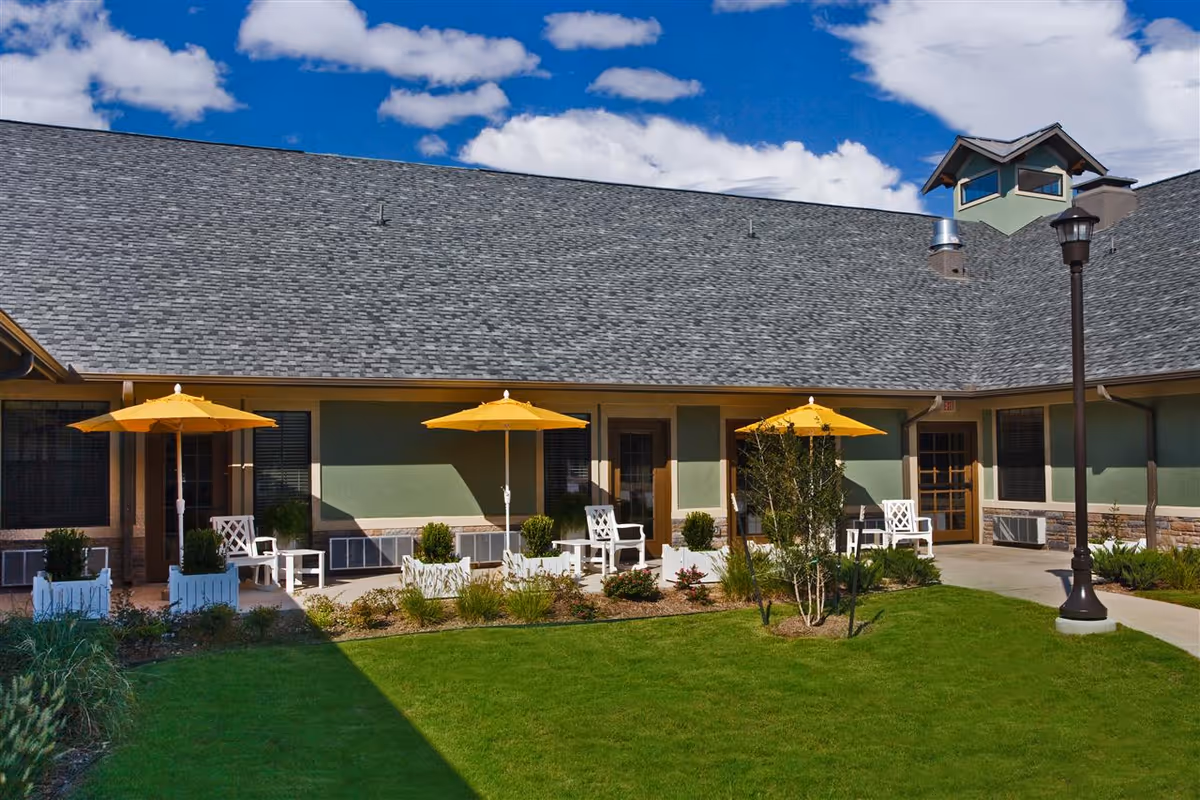 Outdoor patio area at The Belmont at Twin Creeks with green grass, yellow umbrellas, white chairs, and small tables in front of a building with green walls and a gray roof under a partly cloudy blue sky.