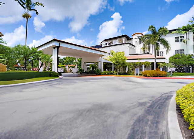 Exterior view of Covenant Village Care Center showing a large white building with multiple windows, a covered driveway entrance, palm trees, and well-maintained landscaping under a partly cloudy blue sky.
