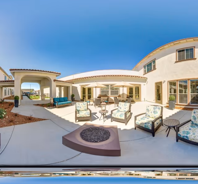 Sunny outdoor courtyard with patio seating, umbrellas, and a central fire pit in front of a two-story building.