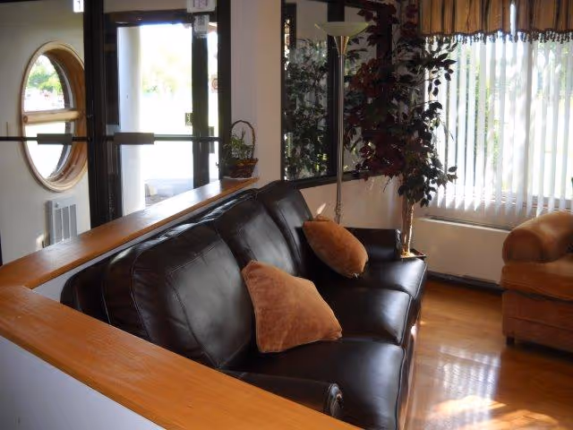 Interior view of a living room area with a black leather couch adorned with two brown pillows, a floor lamp, a large window with vertical blinds, a potted plant, and a brown armchair. There is a wooden railing behind the couch and a round window near the entrance door.