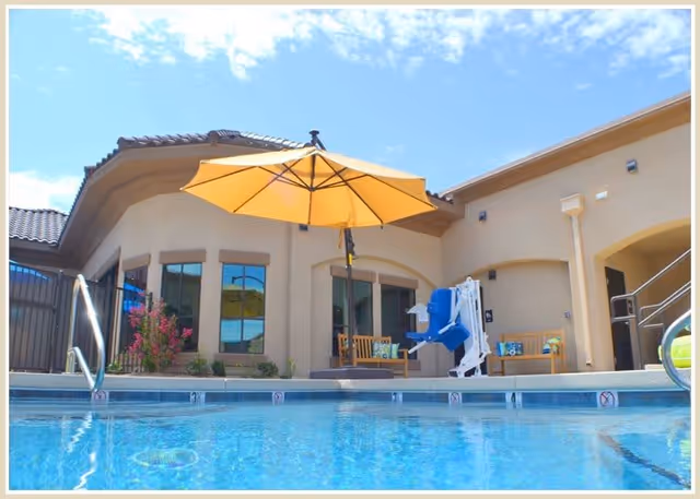 Outdoor swimming pool area at Spanish Hills Wellness Suites with a large yellow umbrella, pool lift chair, and seating benches against the building under a clear blue sky.