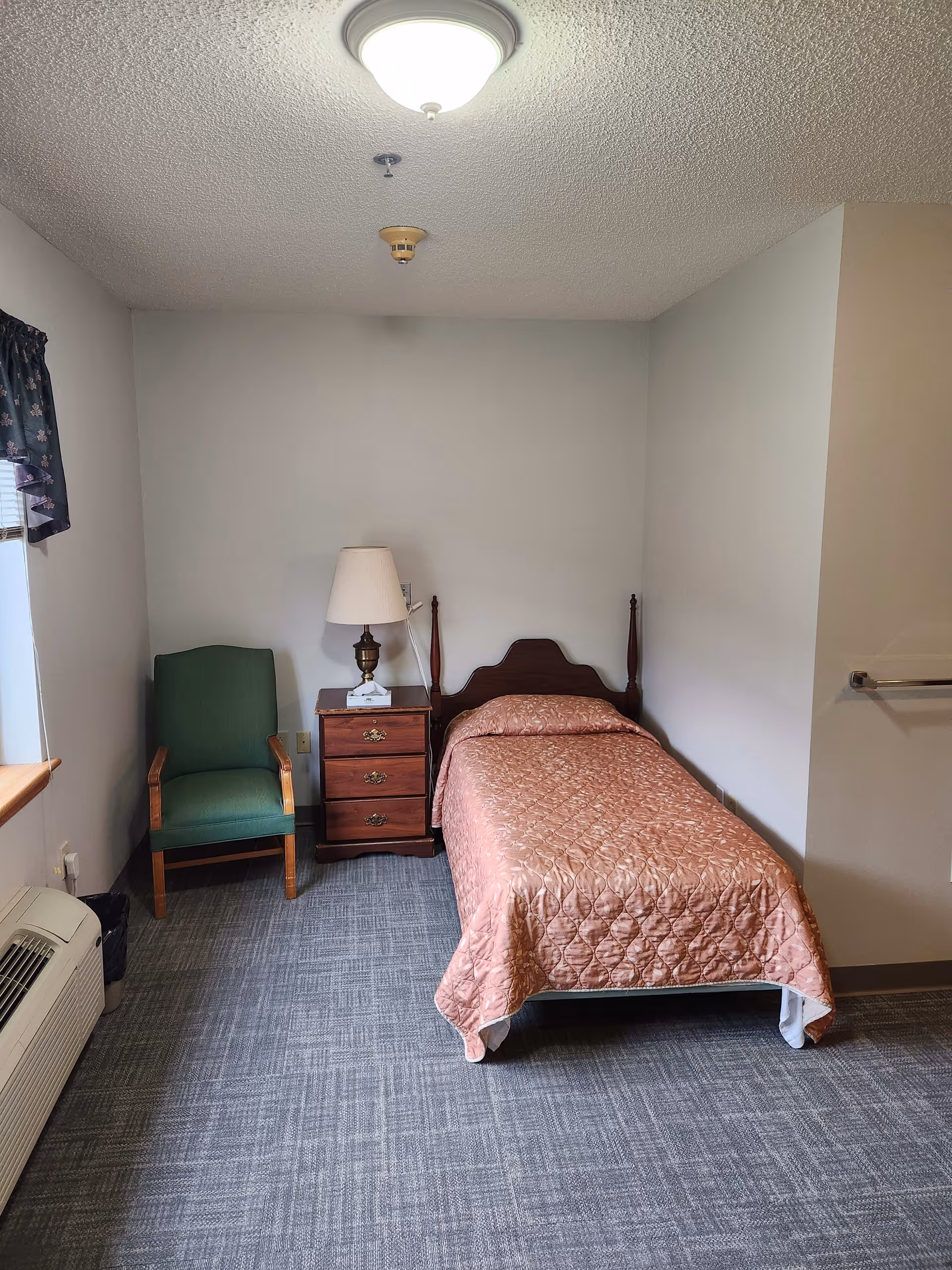 A small, simple bedroom with a single bed covered in a pink quilted bedspread. Next to the bed is a wooden nightstand with three drawers and a lamp on top. To the left of the nightstand is a green upholstered chair. The room has gray carpet flooring, white walls, a window with a floral valance, and a ceiling light fixture.