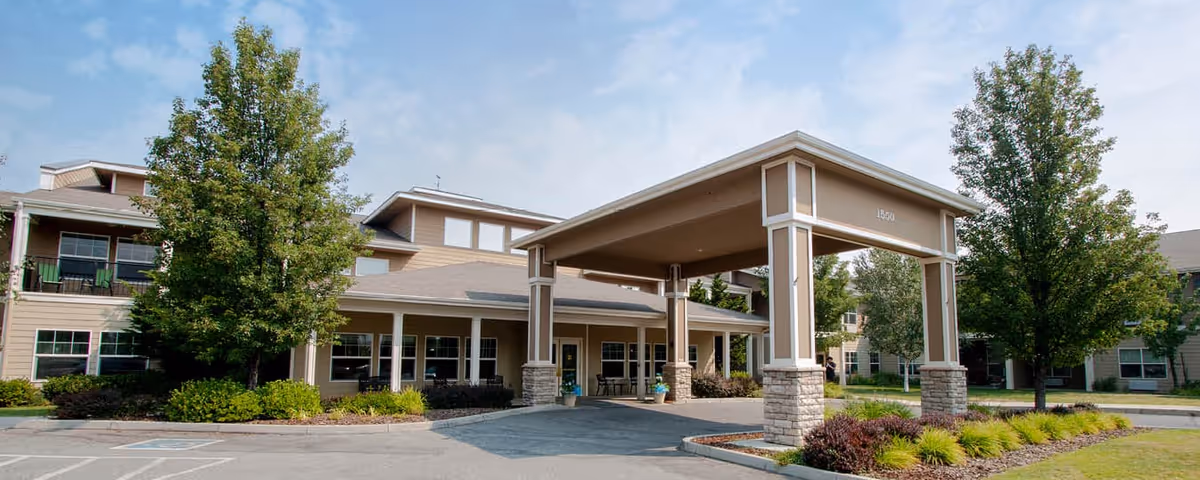 Exterior view of Wenatchee Senior Living by Cogir showing the entrance with a covered drop-off area, surrounded by trees and landscaping under a partly cloudy sky.