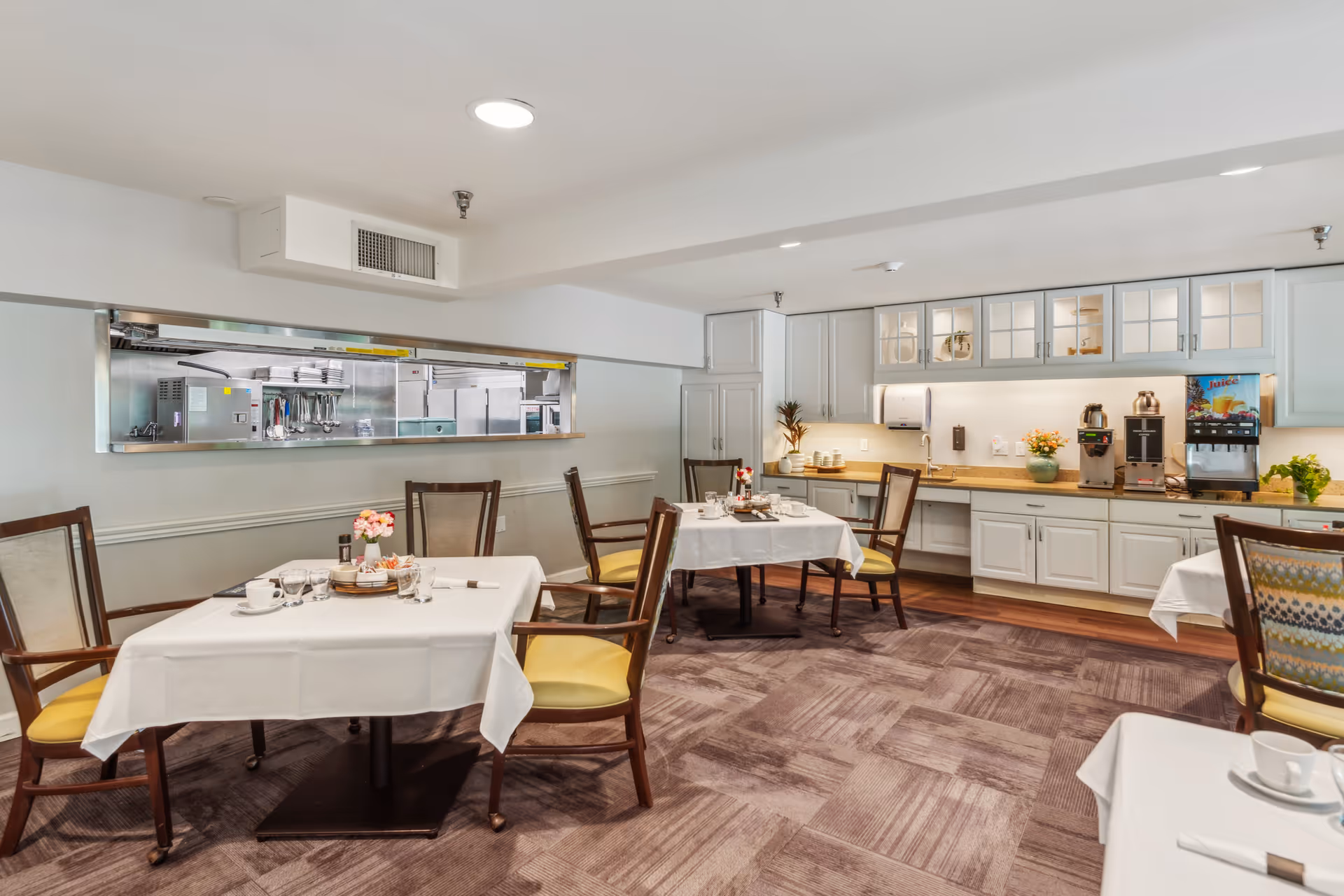 A bright dining room with several set tables and chairs, a serving counter and a kitchen pass-through window.