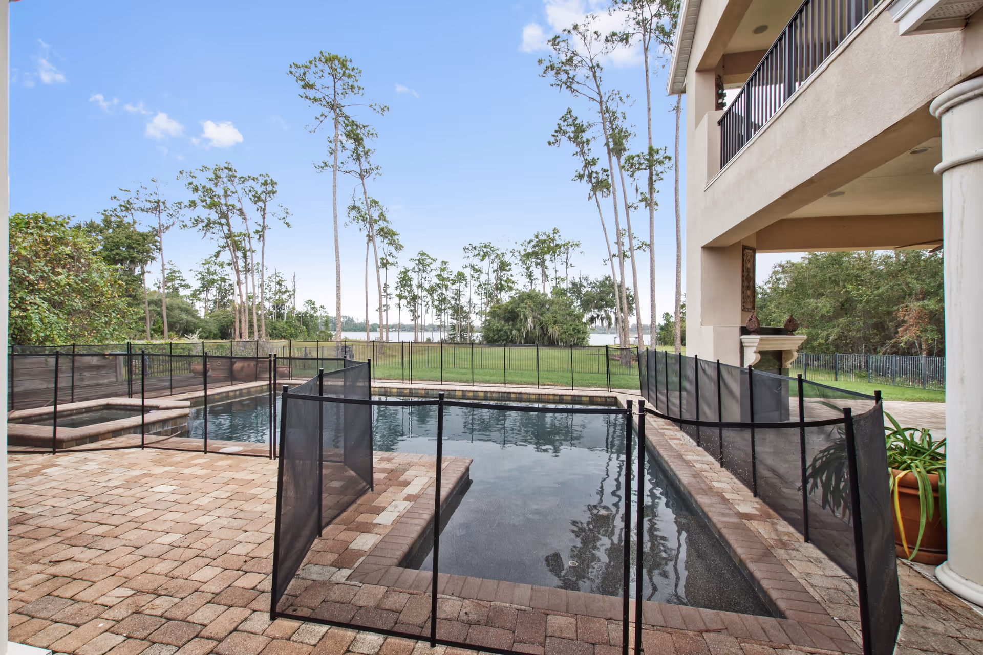Outdoor swimming pool area with a safety fence around the pool, surrounded by a paved patio. Tall trees and a grassy area are visible in the background under a clear blue sky. Part of a building with columns and a balcony is visible on the right side.
