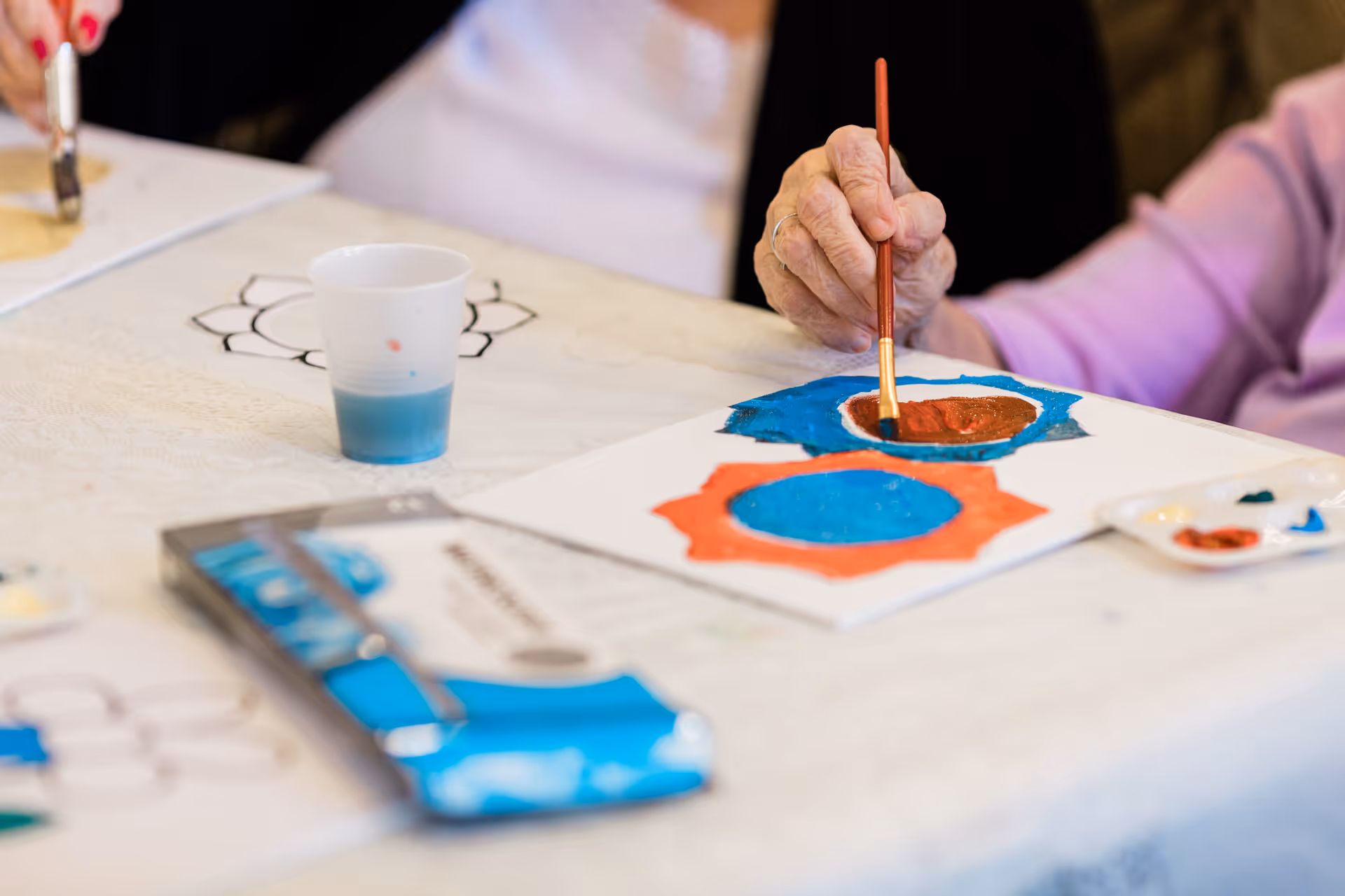 Close-up of elderly hands painting colorful shapes on paper at a table, with a small cup of blue paint and a paint palette nearby.
