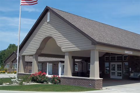 Front exterior view of a senior living facility building with a covered entrance supported by columns, an American flag on a flagpole, and landscaping with green grass and flowering bushes under a clear blue sky.