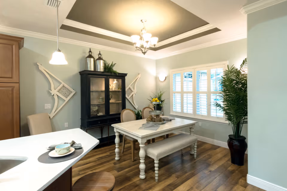 A bright dining room featuring a white wooden dining table with a matching bench and two chairs. The room has light green walls, wooden flooring, and a large window with white shutters. A black cabinet with decorative items is against the wall, flanked by two white wall decorations. A potted plant is in the corner, and the ceiling has a chandelier and recessed lighting. Part of a kitchen counter with a place setting is visible in the foreground.