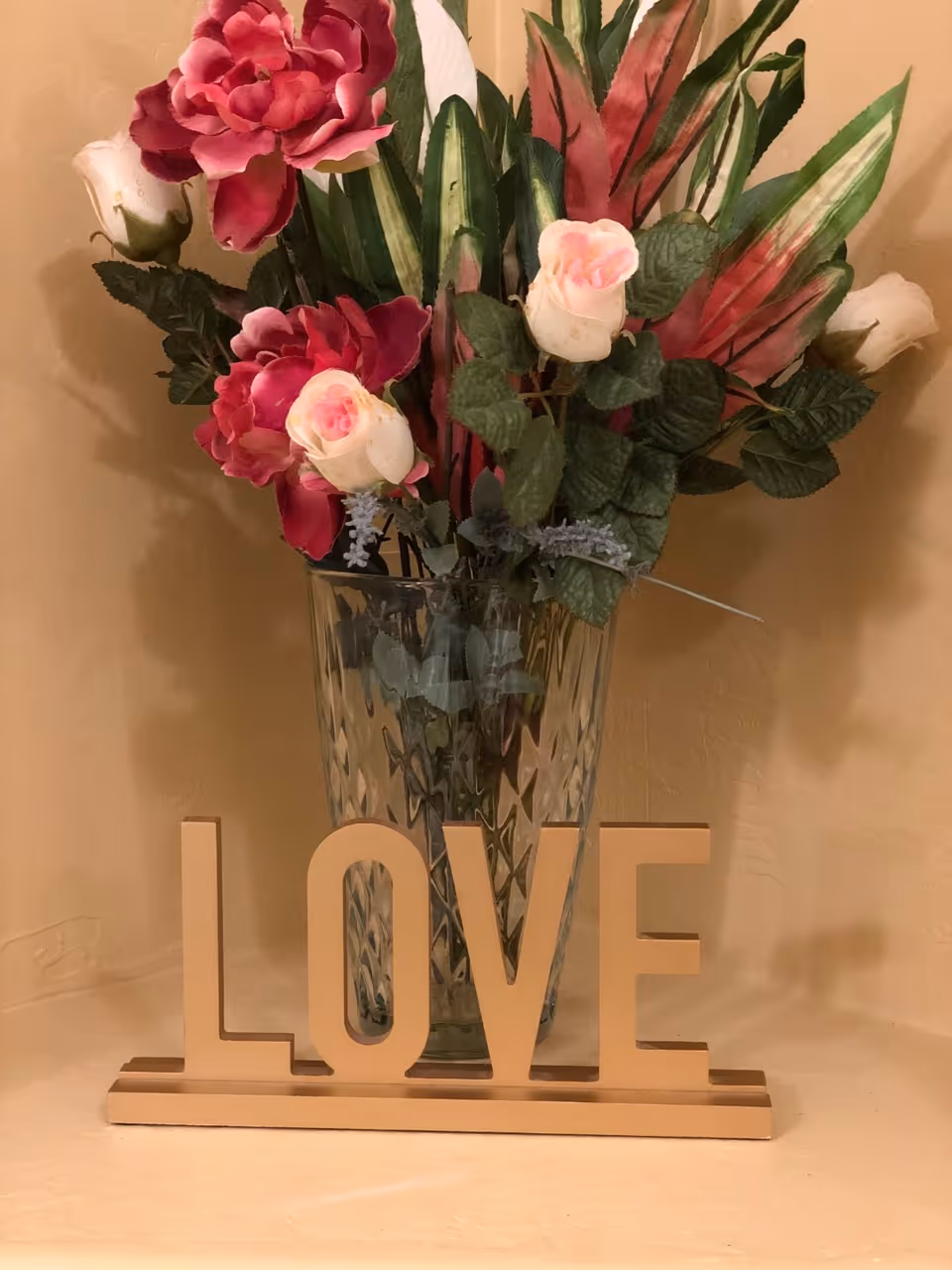 Vase of pink and white artificial flowers behind a wooden 'LOVE' sign on a shelf against a beige wall.