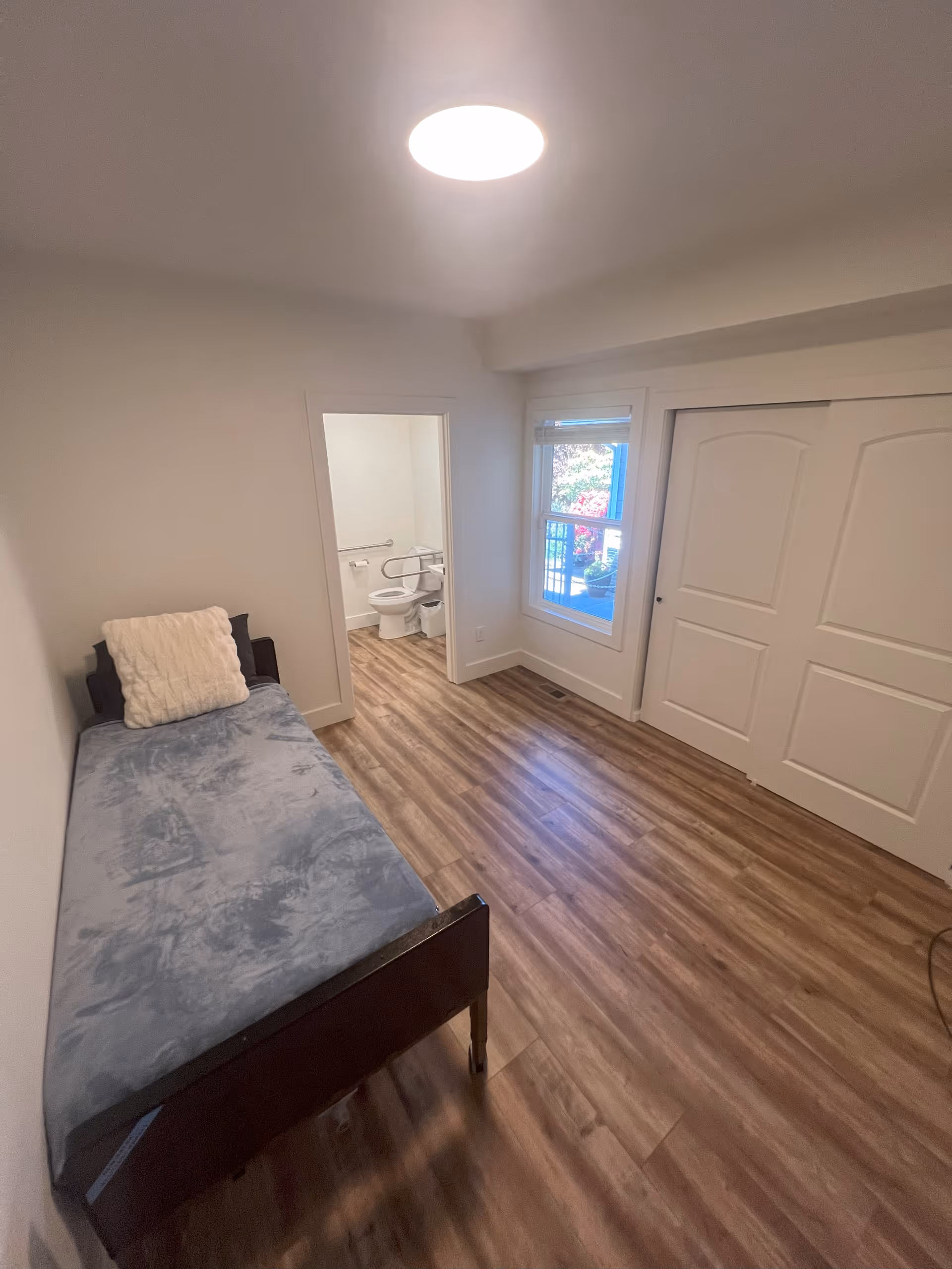 A small single bedroom with a twin bed, wood-look flooring, a window and closet, and an open doorway leading to a bathroom.