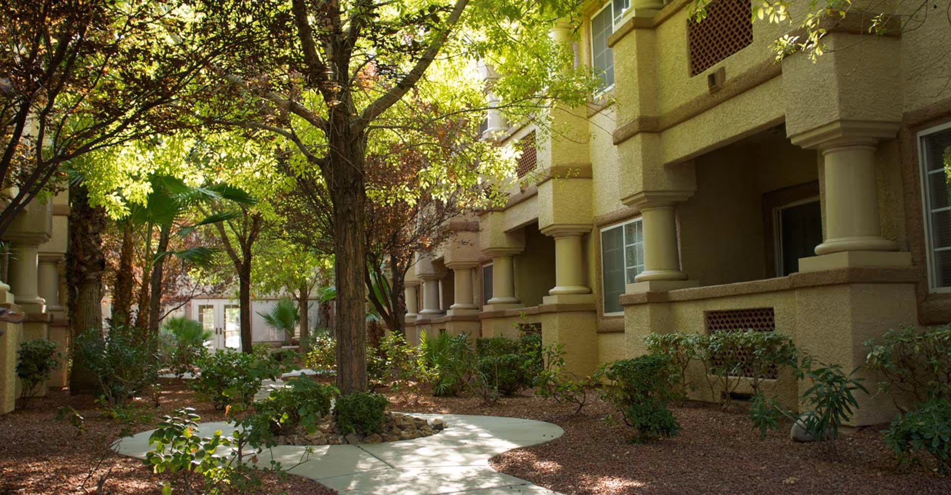 Shaded courtyard with a winding walkway, trees, and landscaping between beige assisted-living building facades.