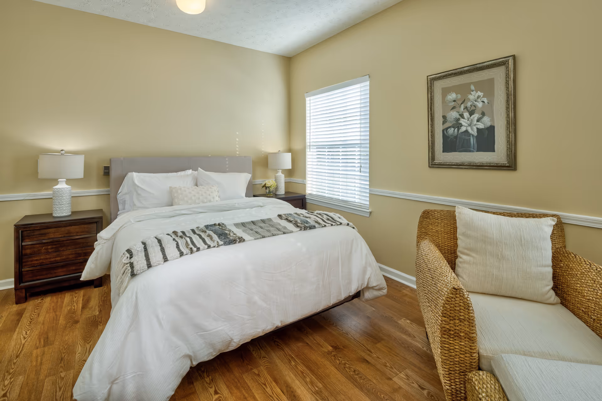 A cozy bedroom with a neatly made bed featuring white bedding and a decorative throw. There are two wooden nightstands with white lamps on either side of the bed. A window with white blinds allows natural light into the room. A wicker armchair with a white cushion is positioned near the window, and a framed floral artwork hangs on the wall above the chair. The room has light yellow walls and wooden flooring.