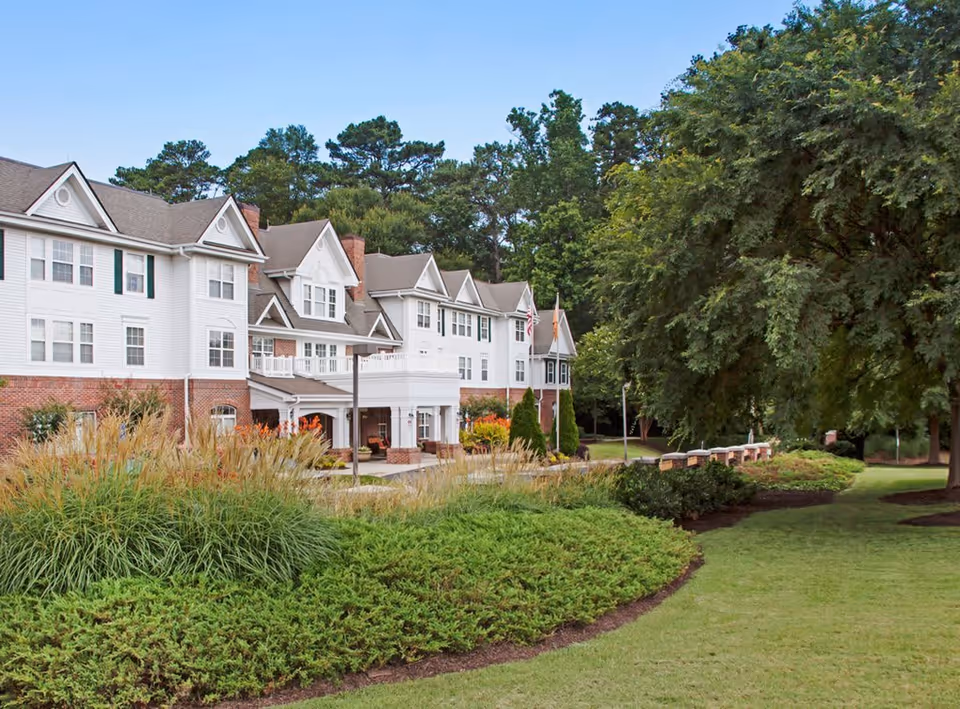 Exterior view of PruittPlace - Buckhead, a multi-story senior living facility with white siding and brick accents, surrounded by well-maintained landscaping including green bushes, tall grasses, and large trees under a clear blue sky.